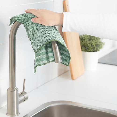 Persona sosteniendo un paño de limpieza verde debajo de un grifo de cocina, con una tabla de cortar y una planta en el fondo.