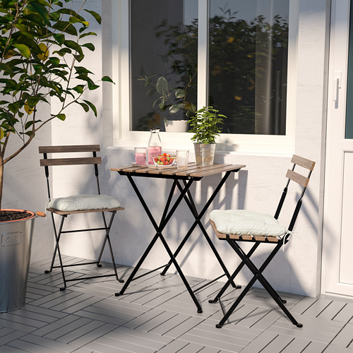 Pequeño bistró TÄRNÖ de metal negro con encimeras de madera de acacia y cojines crema en un balcón, junto a una pared blanca con una ventana y plantas en macetas.