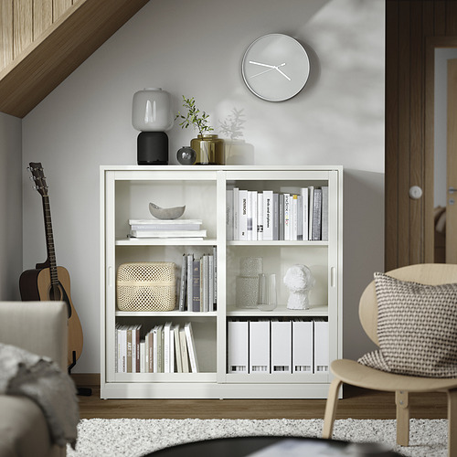 Salón minimalista con estantería blanca TONSTAD, con puertas correderas, jarrones de cristal, decoración apilada y reloj de pared.