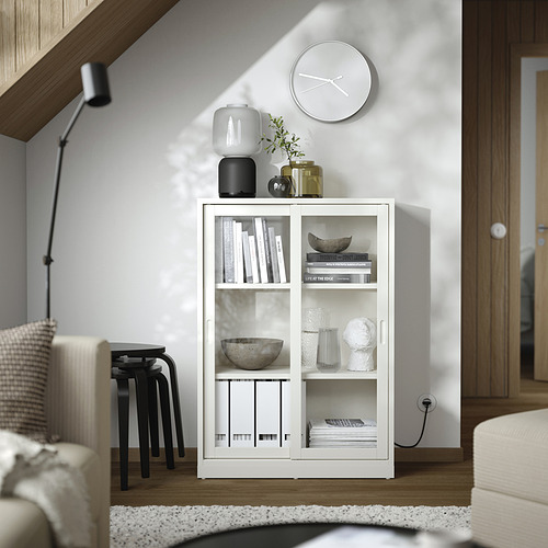 Librería de madera blanca con puertas correderas de vidrio; sostiene libros, jarrones y un reloj, en una sala de estar moderna.