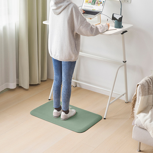 Una persona usando una alfombra de pie verde reciclada FREIVID en una oficina con una silla y una manta cercanas.