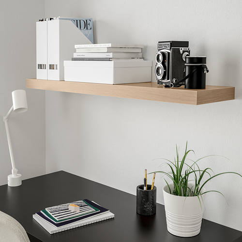 Escritorio moderno con estante de pared blanco LACK que sostiene libros, cámara y planta. Escritorio NEGRO con lámpara, cuaderno y portalápices.