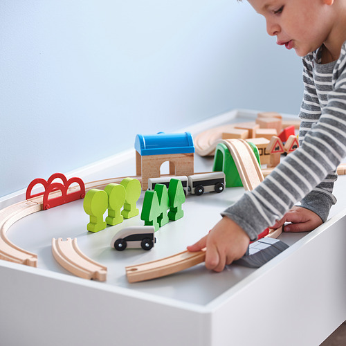 Un niño juega con un tren de madera, arreglando vías, edificios y árboles, demostrando habilidades motoras finas e imaginación.