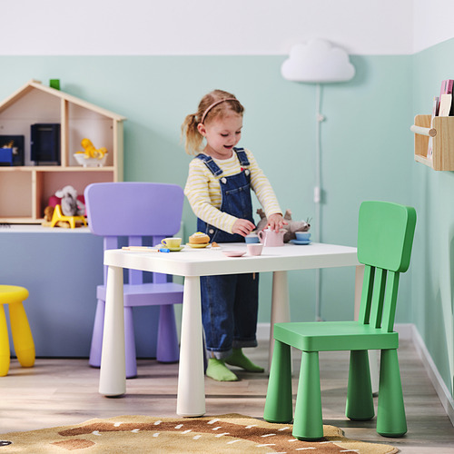 Una niña juega con un juego de té de juguete en una mesa infantil blanca de MAMMUT, rodeada de sillas de colores en una habitación acogedora y llena de luz.
