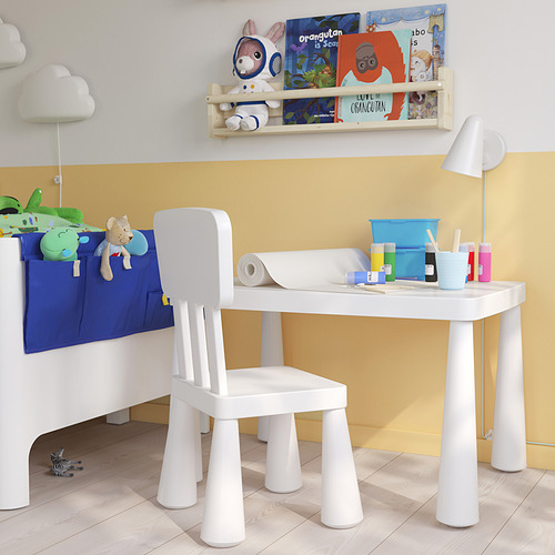 Pequeño juego de mesa y silla blanca para niños con bolsillos de almacenamiento, colocados debajo de un estante de libros coloridos. Ideal para juegos y manualidades.