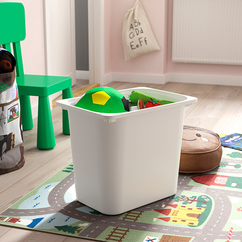 El contenedor de almacenamiento de plástico blanco lleno de bloques verdes y amarillos se encuentra en una alfombra de juego colorida junto a una pequeña silla verde.