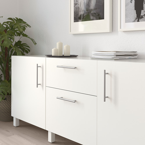 Mueble blanco con tiradores metálicos, dos cajones y dos puertas. Decorado con velas y libros, junto a una planta.