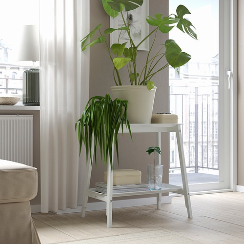 Soporte de plantas cerca de la ventana con una planta grande y frondosa y una pequeña planta con jarrón azul, libro, caja beige.