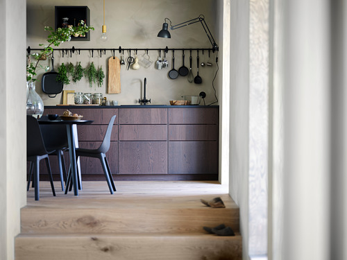 Cocina moderna con armarios de color marrón oscuro, utensilios para colgar y una pequeña mesa de comedor con sillas. Suelo de madera clara y decoración minimalista.