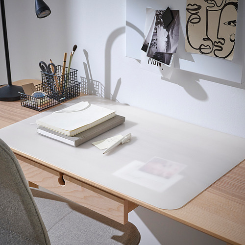 Escritorio ordenado con almohadilla blanca PLÖJA que protege la superficie de madera clara. Cuadernos, bolígrafos e impresiones artísticas en las paredes. Silla gris y lámpara cerca.