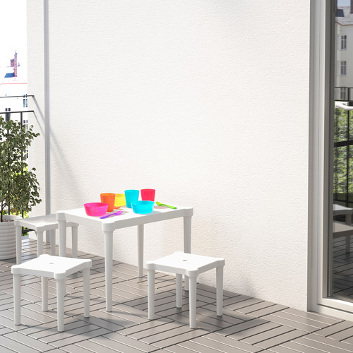 Pequeña mesa blanca para niños y taburetes en el balcón con tazas de colores, contra una pared gris claro.