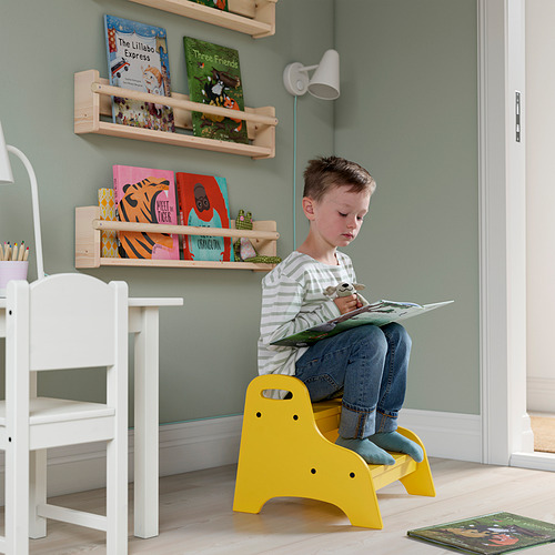 Niño sentado en un taburete amarillo TROGEN, leyendo un libro. Detrás de él, una pared con estanterías de madera llenas de coloridos libros infantiles.