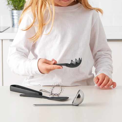 Un niño sosteniendo un servidor de espagueti de juguete, con utensilios de cocina de juguete adicionales en la mesa, promoviendo el juego de cocina de simulación.