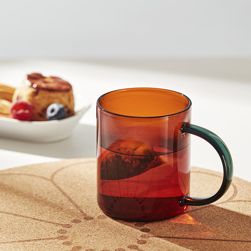 Una taza de vidrio rojo con un mango verde se encuentra sobre una alfombrilla de corcho, mostrando su cómodo agarre y su elegante diseño.
