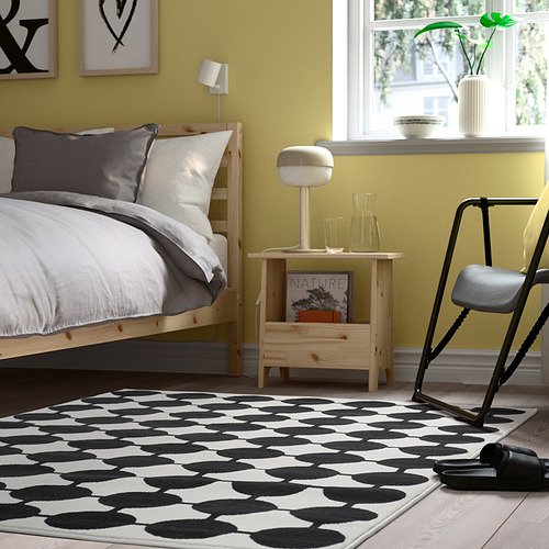 Un dormitorio moderno con una alfombra VÄNDPLATS en blanco y negro, cama de madera, ropa de cama gris/blanca, pared amarilla y decoración minimalista.