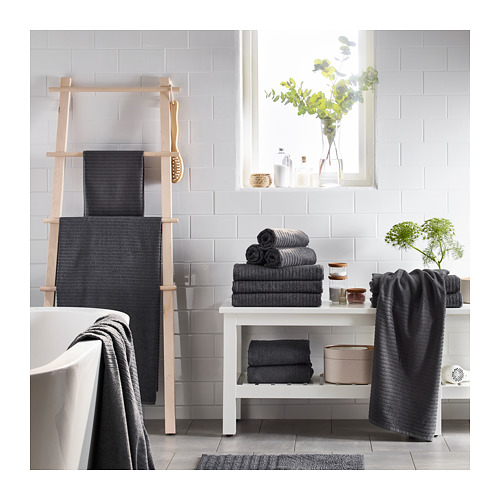 Baño moderno con toallas VÅGSJÖN gris oscuro sobre escalera de madera y mesa blanca, plantas verdes y bañera blanca.