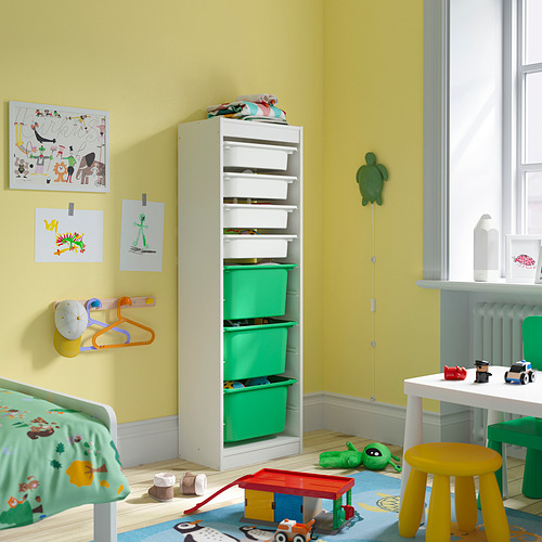 Luminosa habitación infantil con una unidad de almacenamiento TROFAST alta, marco blanco y cajas verdes, con varios juguetes y arte colorido en paredes de color amarillo claro.
