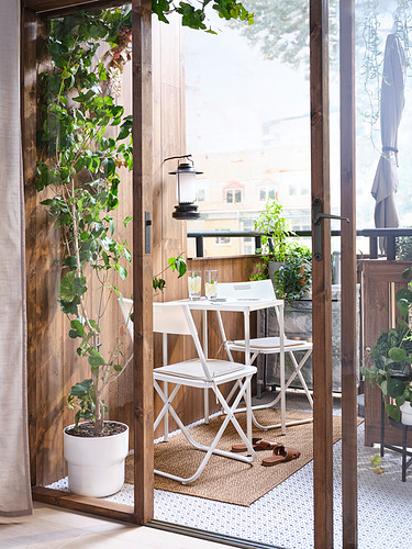 Pequeño balcón con sillas plegables blancas, mesa y plantas en macetas. Suelo de madera, puerta de cristal.