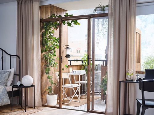 Dormitorio luminoso y moderno con grandes puertas correderas al balcón, con mesa y sillas blancas al aire libre, plantas exuberantes y cortinas transparentes.