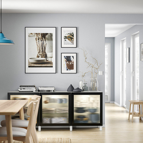 Comedor moderno con mesa y sillas de madera clara, aparador negro y transparente, pared gris, tres fotos enmarcadas, lámpara azul.