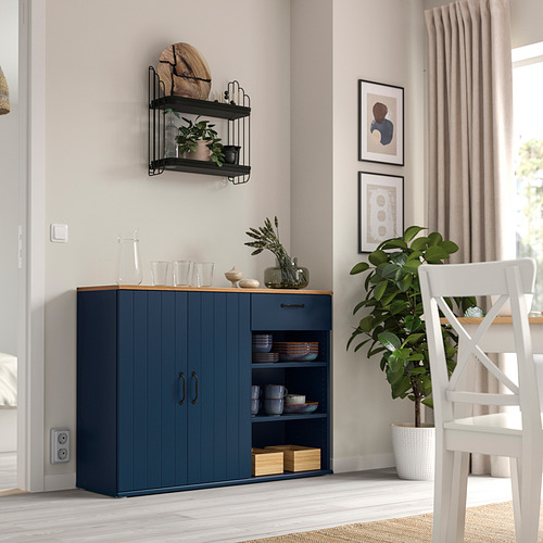 Comedor moderno con aparador SKRUVBY azul y estantes abiertos, complementado por una silla blanca y una gran planta verde, con arte en la pared y cortinas.