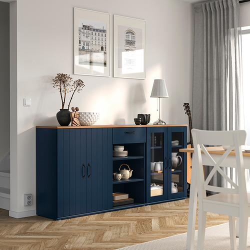 Comedor moderno con aparador SKRUVBY azul, puertas de vidrio y porcelana. Cortinas grises, estampados blancos, suelo de madera.