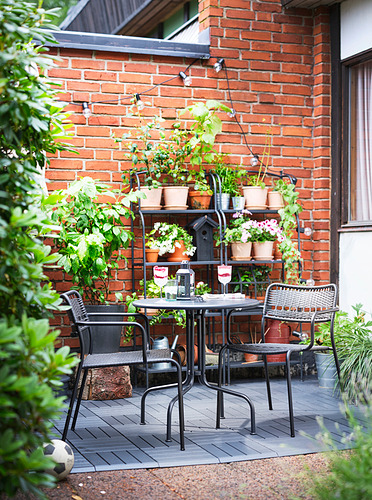 Acogedor patio con muebles de metal negro LÄCKÖ contra pared de ladrillo, plantas y luces.