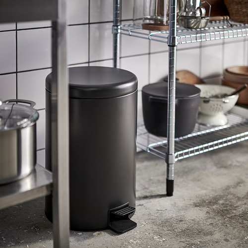 Bote de basura de cocina negro con mecanismo de pedal cerca de estanterías de alambre.