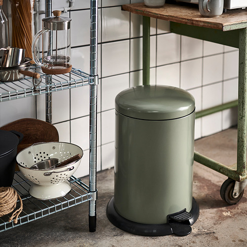 Cubo de basura verde oliva con tapa y pedal en la cocina junto a estantería metálica con artículos.