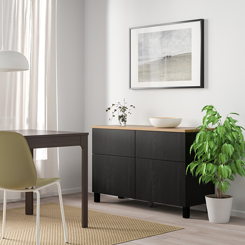 Sala de estar moderna con aparador negro BESTÅ, decoración y silla lima. Luz natural a través de ventana con cortinas blancas. Alfombra beige sobre suelo claro.