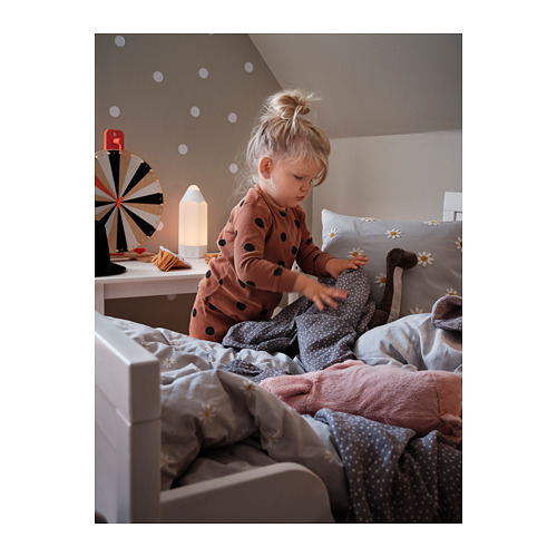 Un niño jugando en una cama con una almohada con estampado de margaritas. La habitación tiene luces de hadas, una lámpara de noche y una colorida peonza en un estante rosa.
