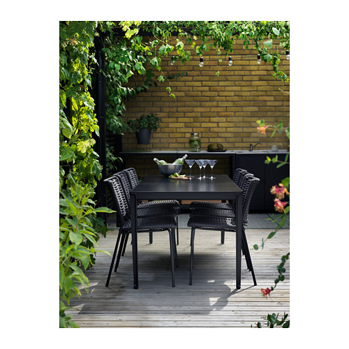 Moderno patio al aire libre con mesa rectangular negra y sillas a juego, rodeado de vegetación y pared de ladrillo.