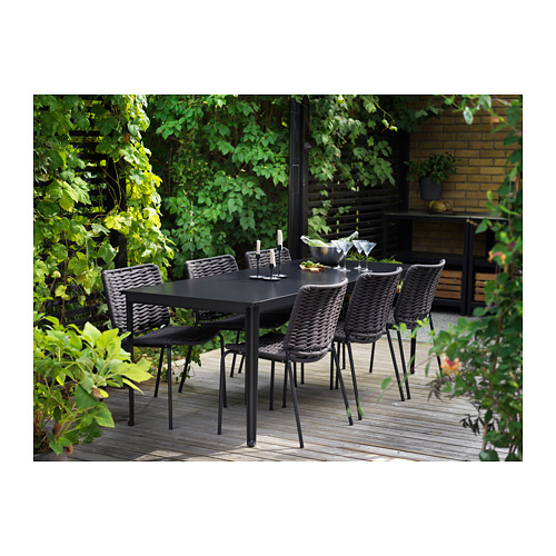 Patio exterior con mesa y sillas TEGELÖN negras, tablero compuesto de madera y plástico y sillas oscuras con asientos de mimbre sintético.