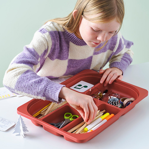 Una niña organiza los materiales de manualidades en una bandeja de almacenamiento TROFAST roja con compartimentos sobre una mesa.