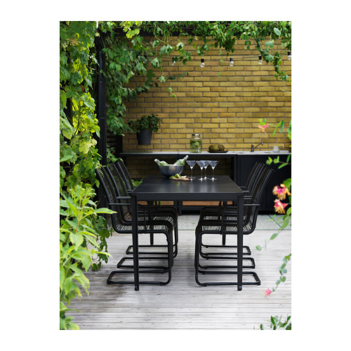 Moderno juego de comedor al aire libre con sillas y mesa negras en una terraza de madera, con un fondo de pared de ladrillo amarillo y exuberante vegetación.