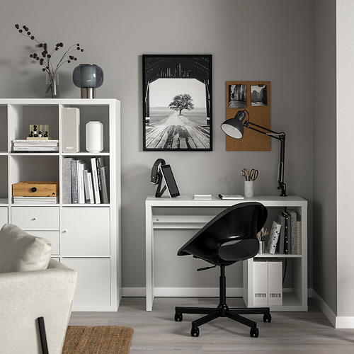 Moderna oficina en casa con escritorio KALLAX, silla giratoria negra, pared gris, estantería, foto, lámpara, tableta y sofá.