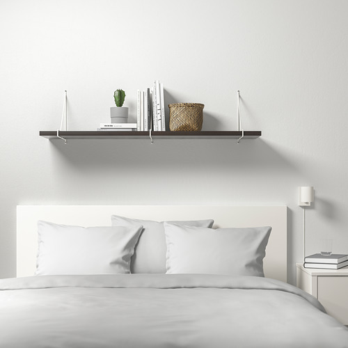 Dormitorio blanco minimalista con estantería de pared BERGSHULT con libros, plantas y cesta, sobre una cama cuidadosamente hecha con ropa de cama blanca.