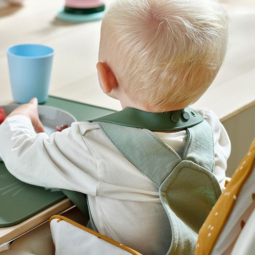 Niño con un babero GULDVÄVARE verde y blanco, diseñado con un bolsillo para atrapar comida, sentado en una silla alta, comiendo.