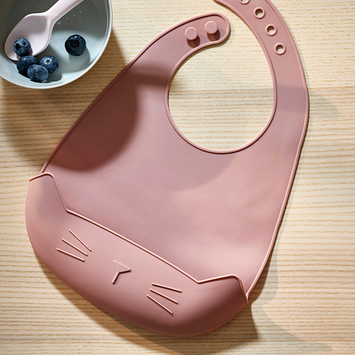 Babero de silicona con forma de gato sobre una mesa de madera, con un bol de arándanos y una cuchara. Cierre ajustable en el cuello, con un bolsillo para atrapar alimentos.