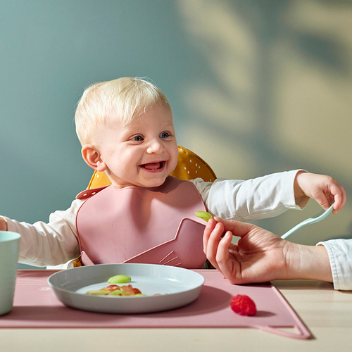 Bebé con un babero de silicona con un bolsillo para atrapar la comida que se cae. Un adulto alimenta al bebé con una cuchara. Un cuenco con fruta está sobre la mesa.