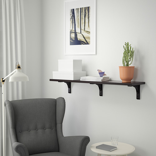 Explora una estantería de pared BERGSHULT, negra, con un borde biselado y otro liso, que exhibe libros y una planta junto a una silla gris y una mesa pequeña.