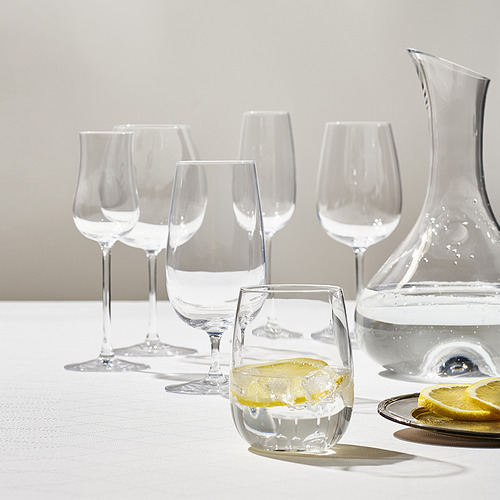 Cinco vasos de cristal STORSINT transparentes y elegantes y una jarra de agua sobre un mantel blanco. Un vaso contiene rodajas de limón.