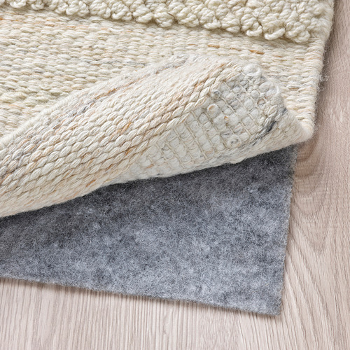 Una alfombra de lana tejida a mano con un patrón texturizado, colocada sobre un suelo de madera con una capa de alfombra de fieltro gris para mayor agarre y comodidad.