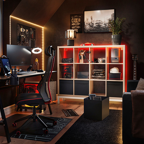 Oficina moderna con estantes KALLAX, detalles en negro y rojo, piso de madera, paredes oscuras, configuración de computadora, guitarra y artículos decorativos.
