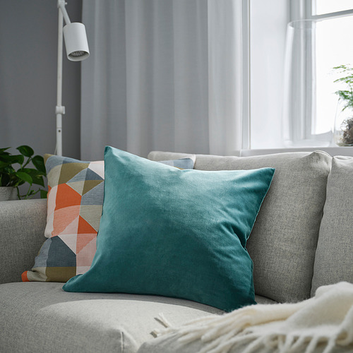 Moderno salón con un sofá gris, un gran cojín de terciopelo turquesa SANELA y una almohada estampada contra una ventana con cortinas ligeras.