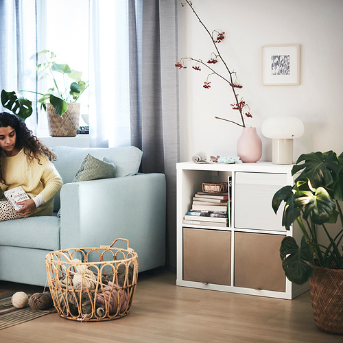 Acogedora sala de estar: mujer lee en sofá azul, estantes KALLAX con libros y plantas, cesta de lana, luz natural.