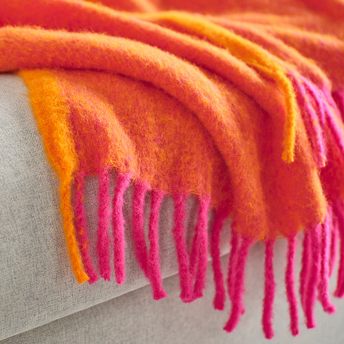 Manta rosa brillante y naranja con textura suave y esponjosa, colocada sobre el borde de los muebles, mostrando extremos con flecos.