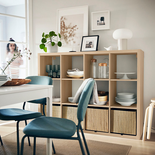 Sala de estar moderna con estante de roble KALLAX, sillas azules, decoración blanca, plantas y arte enmarcado.