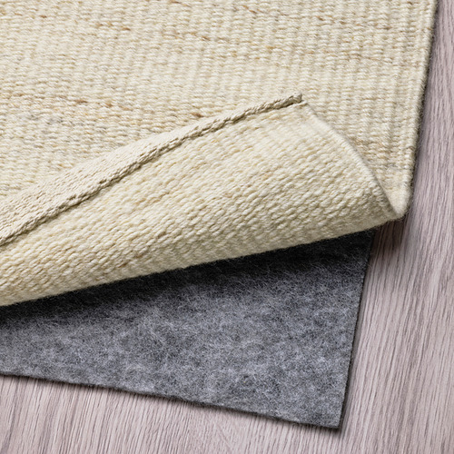 Una alfombra de lana beige doblada, dejando al descubierto su parte inferior. Una alfombra de fieltro gris debajo cubre la parte inferior, evitando resbalones.