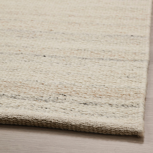 Alfombra beige de tejido plano con textura sutil, colocada sobre suelo de madera. Los materiales de calidad dan una sensación suave y cálida.